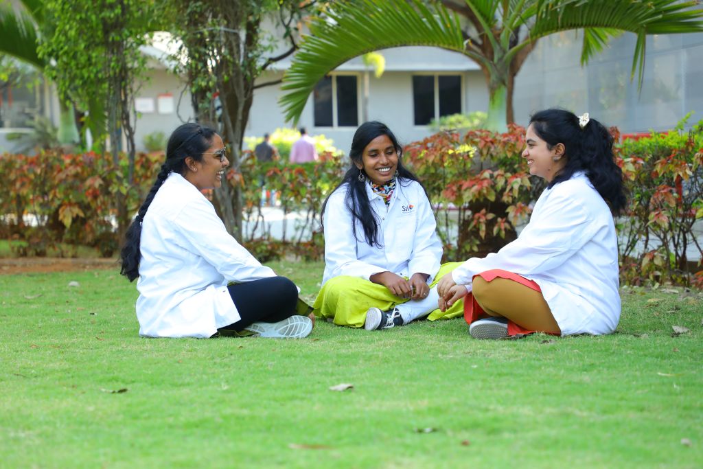 Three female scientists wearing white lab coats sit on a green lawn, engaged in a friendly conversation, outside a modern research facility with lush greenery in the background.