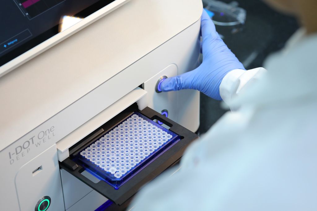 Scientist wearing blue gloves operating an I-DOT One Deep Well liquid handling system, preparing a 96-well plate for high-throughput screening in a drug discovery laboratory.