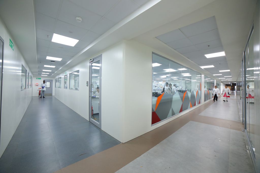 A modern laboratory facility at Sai Life Sciences with a clean, well-lit corridor and glass-walled research labs. Scientists in white lab coats can be seen working inside, reflecting a high-tech and collaborative research environment.