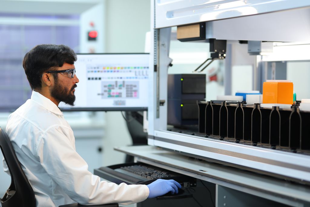 Scientist in a lab coat and safety glasses operating an automated liquid handling system for high-throughput drug metabolism and pharmacokinetics (DMPK) research in a laboratory setting.