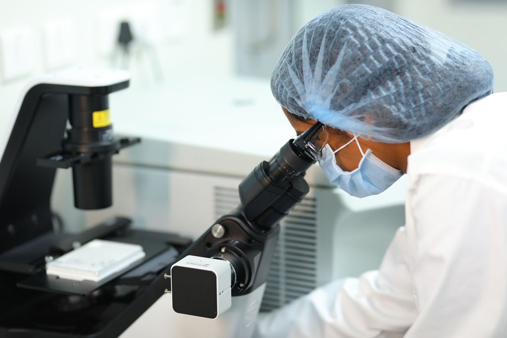 Scientist examining a sample under a high-powered microscope in a pharmaceutical laboratory, wearing protective gear, including a hairnet, face mask, and lab coat.