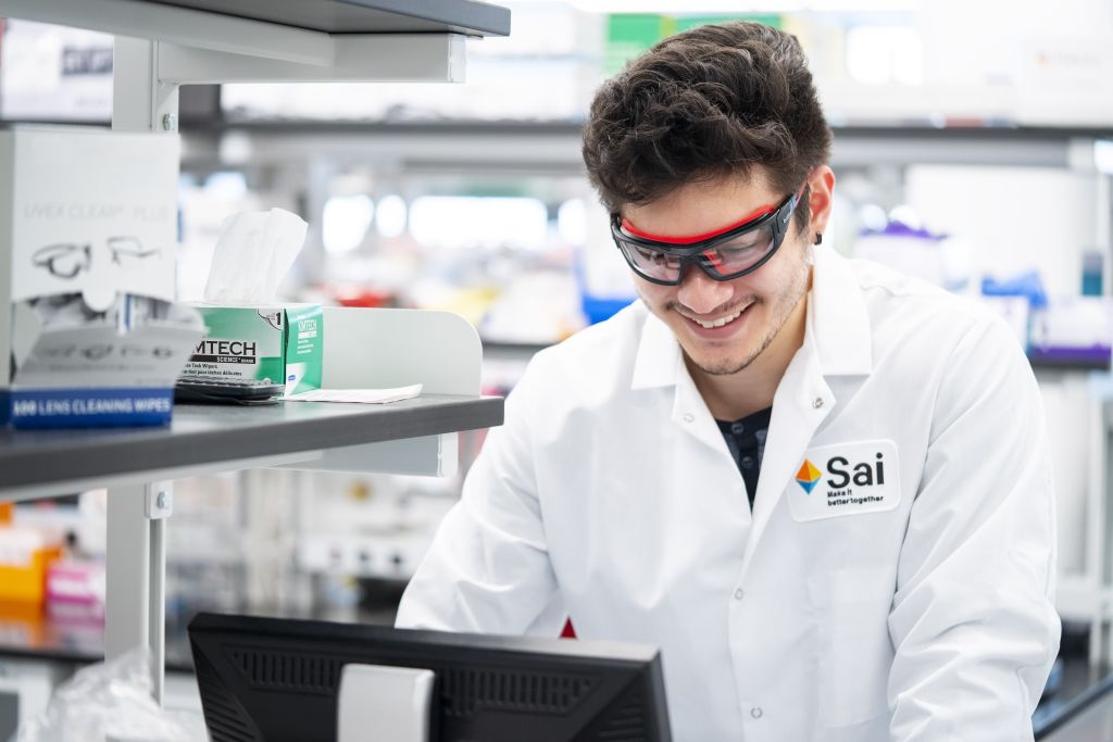 Sai Life Sciences employee wearing safety goggles and a lab coat, smiling while working on a computer in a well-equipped laboratory. Shelves stocked with scientific supplies and equipment are visible in the background. The employee badge displays the company logo with the tagline 'Make It Better Together.'