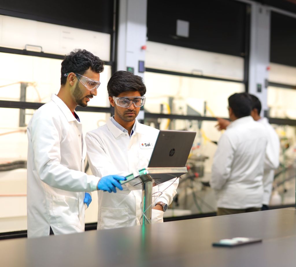 Sai Life Sciences scientists collaborating in a laboratory, wearing safety goggles and lab coats, analyzing data on a laptop, with a background of chemical fume hoods and additional researchers